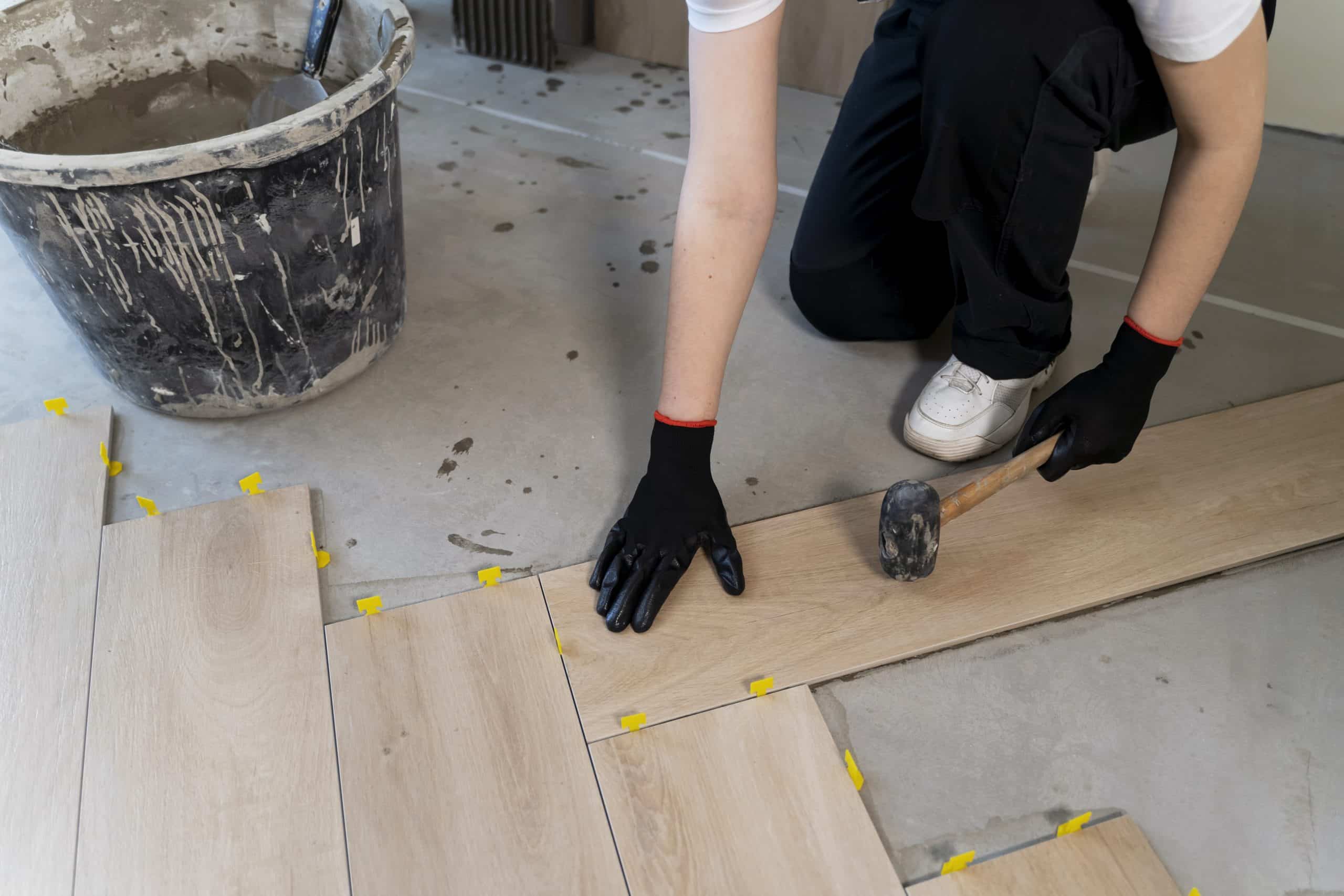 Eine Person in schwarzer Kleidung und Handschuhen verlegt Holzpaneele auf einem Boden. Sie hält einen Gummihammer in der rechten Hand und hat die linke Hand auf einem Paneel platziert. Im Hintergrund steht ein Eimer mit Mörtel und gelbe Abstandshalter sind sichtbar.