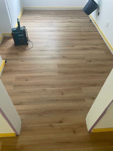 Ein neu verlegter Holzboden in einem Raum, mit einem blauen Werkzeug und einem Abfalleimer im Hintergrund. Die Wände sind gelb gestrichen, und der Boden zeigt eine natürliche Holzmaserung.