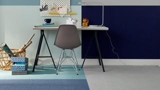 Ein moderner Arbeitsplatz mit einem weißen Schreibtisch, einem grauen Stuhl und bunten Teppichen in verschiedenen Farben. An der Wand hängen organisatorische Elemente und eine Lampe steht auf dem Tisch.