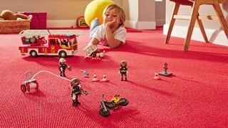 Ein Kind liegt auf einem roten Teppich und spielt mit Spielzeugfiguren, darunter Feuerwehrleute, ein Feuerwehrauto und ein Motorrad. Im Hintergrund sind weitere Spielzeuge und ein Stuhl zu sehen.