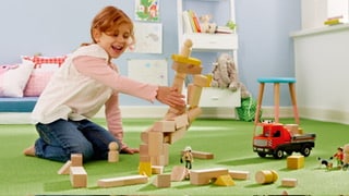 Ein fröhliches Kind spielt auf einem grünen Teppich mit Holzspielzeug und baut mit bunten Bauklötzen. Neben ihr steht ein rotes Spielzeugauto, während sie konzentriert an ihrer Konstruktion arbeitet. Im Hintergrund sind Regale mit Spielzeug und Bildern zu sehen.