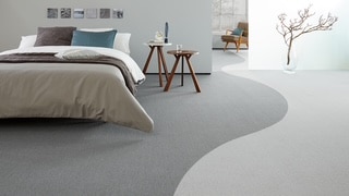 Ein modernes Schlafzimmer mit einem grauen Teppichboden, einem Bett mit einer neutralen Bettdecke und Kissen in verschiedenen Farben. Neben dem Bett steht ein kleiner Tisch, und im Hintergrund sind eine Pflanze und ein Fenster zu sehen.